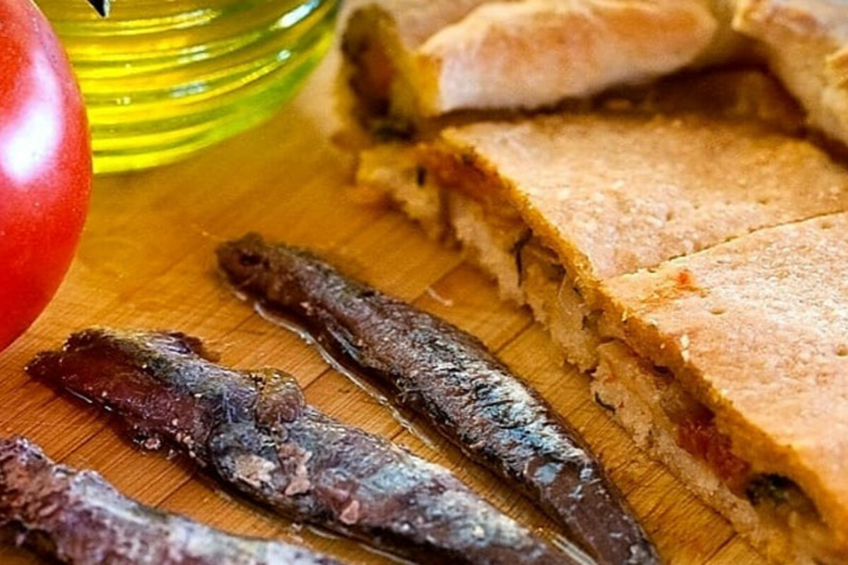 Tradicionalna viška pogača narezana na trokute, poslužena uz slane inćune, s maslinovim uljem i rajčicom u pozadini, na drvenoj podlozi.