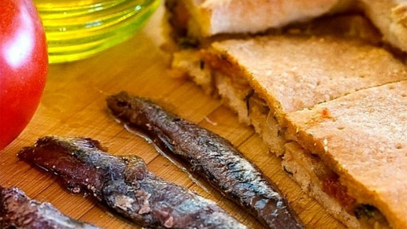 Tradicionalna viška pogača narezana na trokute, poslužena uz slane inćune, s maslinovim uljem i rajčicom u pozadini, na drvenoj podlozi.