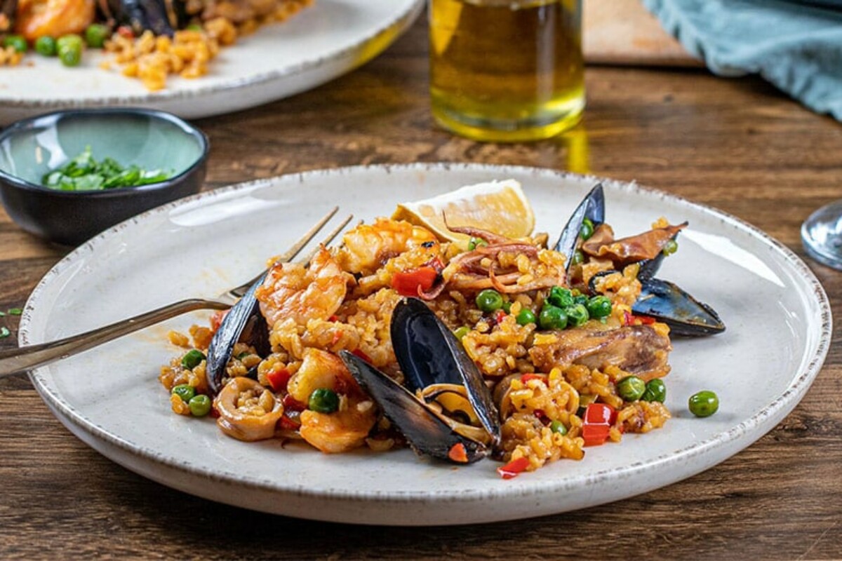 Španjolska paella poslužena na bijelom tanjuru, s rižom, plodovima mora, komadićima piletine, graškom, paprikom i kriškom limuna.