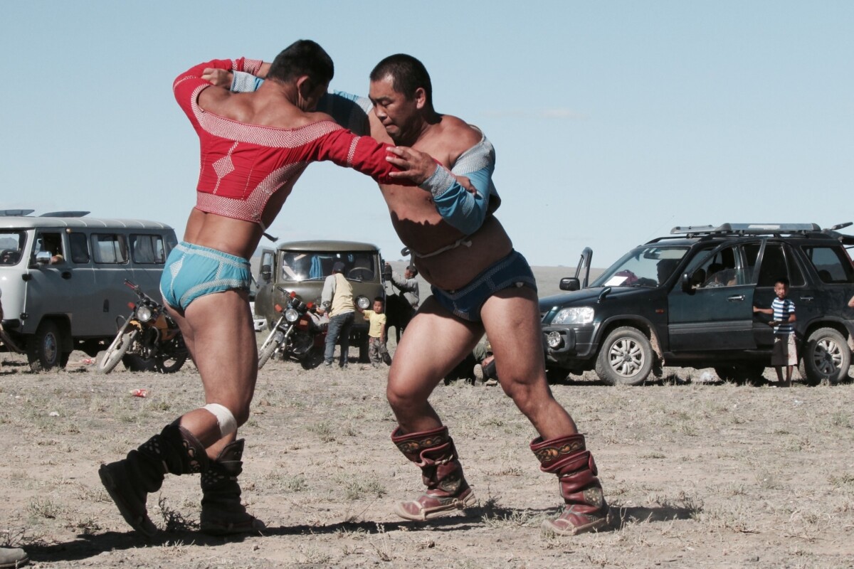 Dvojica mongolskih hrvača u tradicionalnim šarenim kostimima natječu se u hrvanju, jednom od tri glavna sporta festivala Naadam. Natjecanje se odvija na otvorenom, uz prisustvo gledatelja i parkiranih vozila u pozadini, odražavajući autentičnu atmosferu mongolske tradicije.