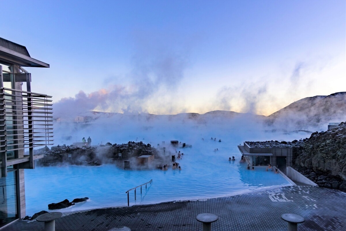 Pogled na Plavu lagunu na Islandu u zoru – ljudi se opuštaju u toplim termalnim vodama okruženim parom i vulkanskim stijenama.