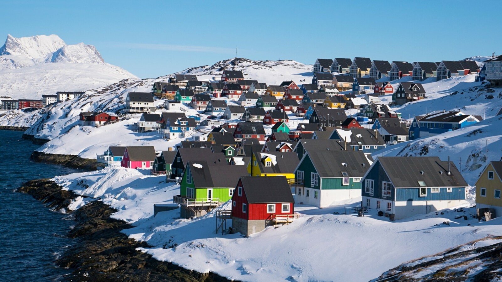 Pogled na Nuuk, glavni grad Grenlanda, s mnoštvom šarenih kuća smještenih uz obalu, prekrivenih snijegom, s morem i planinama u pozadini pod plavim nebom.