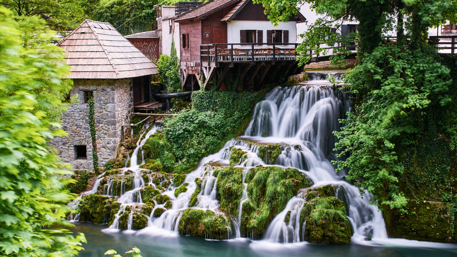 rastoke-slapovi Slapovi u Rastokama, okruženi zelenilom i tradicionalnim drvenim kućama na rijeci Slunjčici, s mlinom od kamena i drvenim mostićem iznad vode.