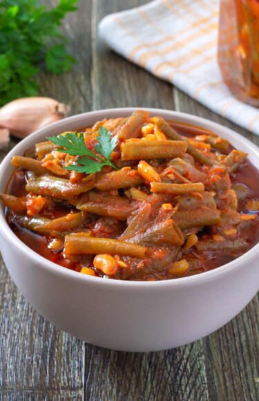 varivo-od-mahuna-recept Varivo od mahuna u umaku od rajčice, posluženo u zdjeli na drvenom stolu s češnjakom, peršinom i ljutom papričicom – ukusno domaće povrtno jelo.