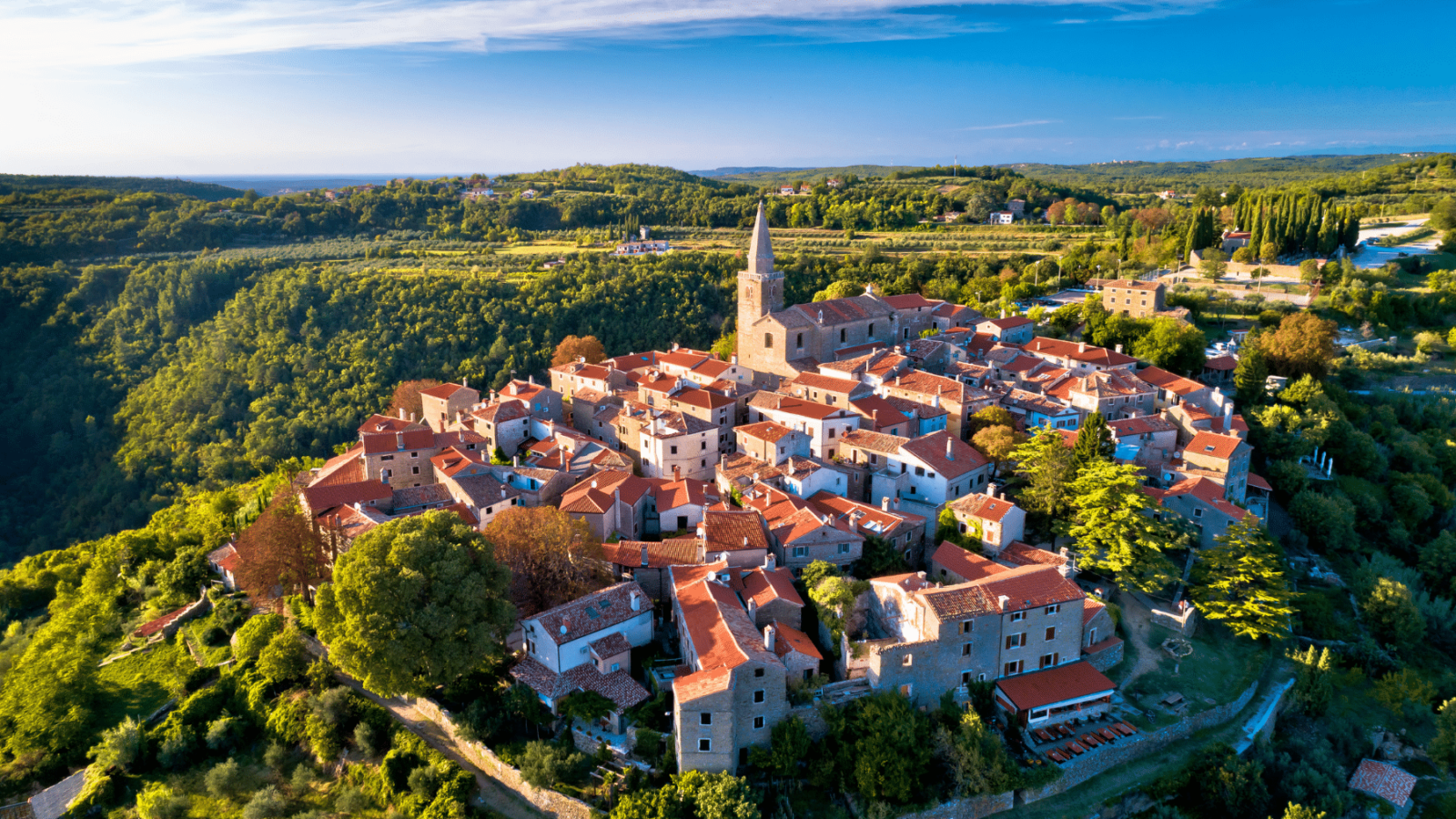groznjan-istra Pogled iz zraka na Grožnjan, šarmantno istarsko naselje s kamenim kućama i crvenim krovovima, smješteno na brežuljku usred zelenog krajolika, s crkvom koja dominira središtem mjesta.