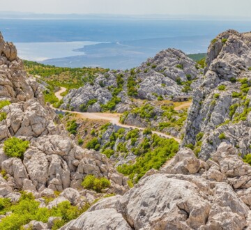 velebit-majstorska-cesta-pogled Pogled na Majstorsku cestu koja vijuga kroz impresivne stijene Velebita, s pogledom na more u daljini i okolicu prekrivenu zelenilom.