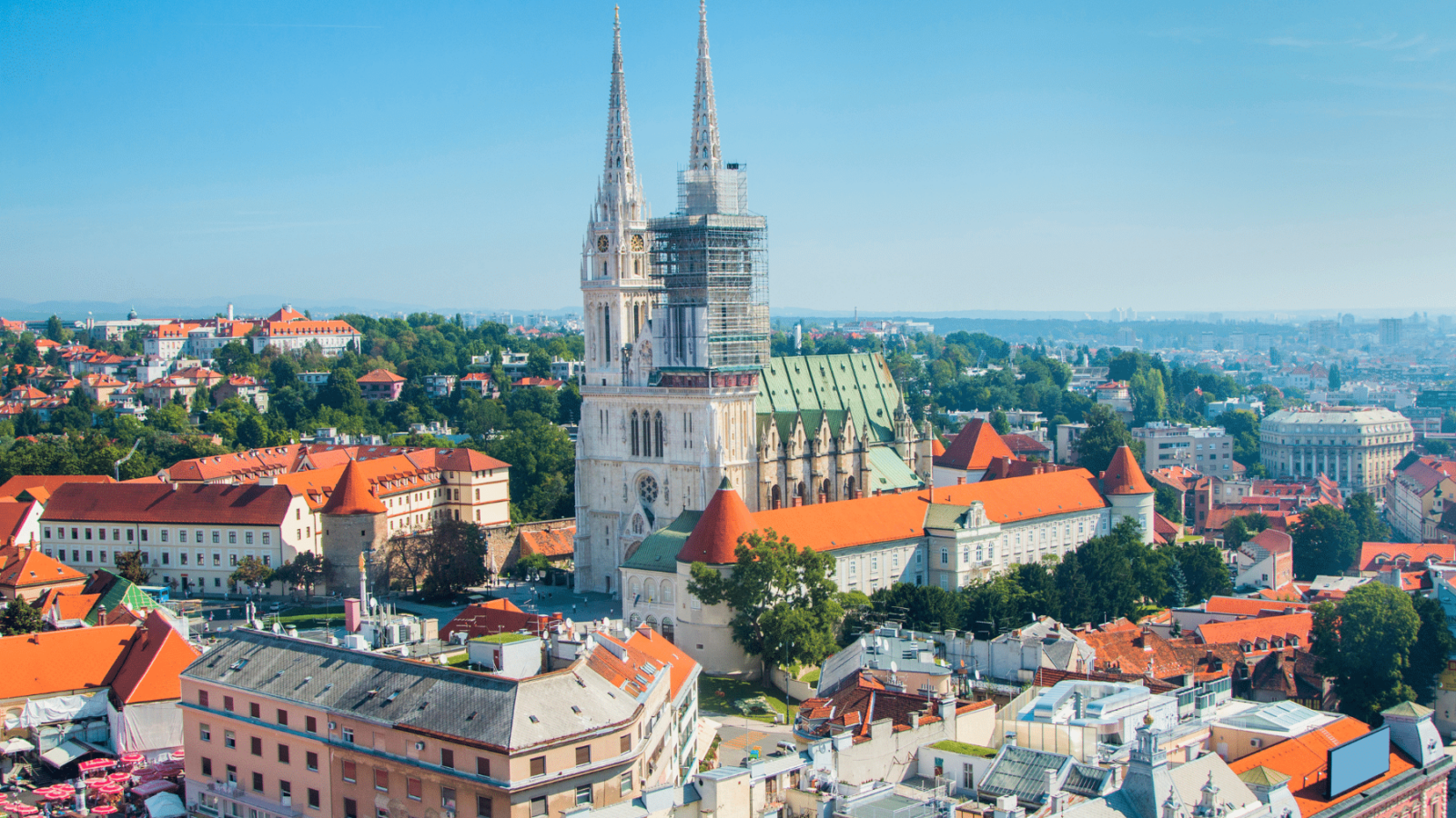 zagreb-katedrala Panoramski pogled na Zagreb s dominantnom zagrebačkom katedralom, čije tornjeve krasi impozantna gotička arhitektura. U prvom planu se nalazi živopisna tržnica Dolac s crvenim suncobranima, dok se u pozadini prostire gradski pejzaž ispunjen crvenim krovovima i zelenim brežuljcima.