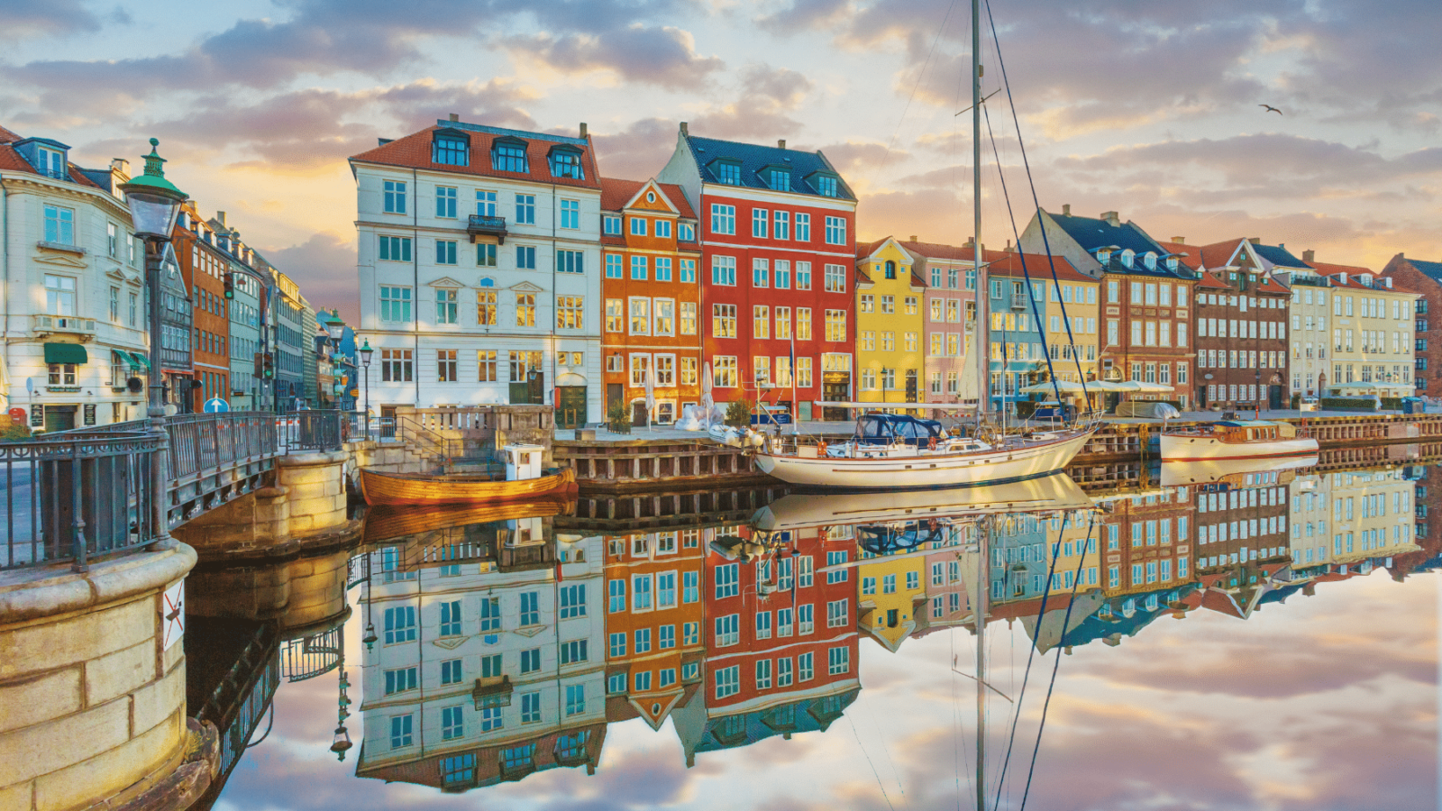 Šarene povijesne kuće uz kanal Nyhavn u Kopenhagenu, s odrazom u mirnoj vodi i brodovima usidrenima duž obale pri zalasku sunca.