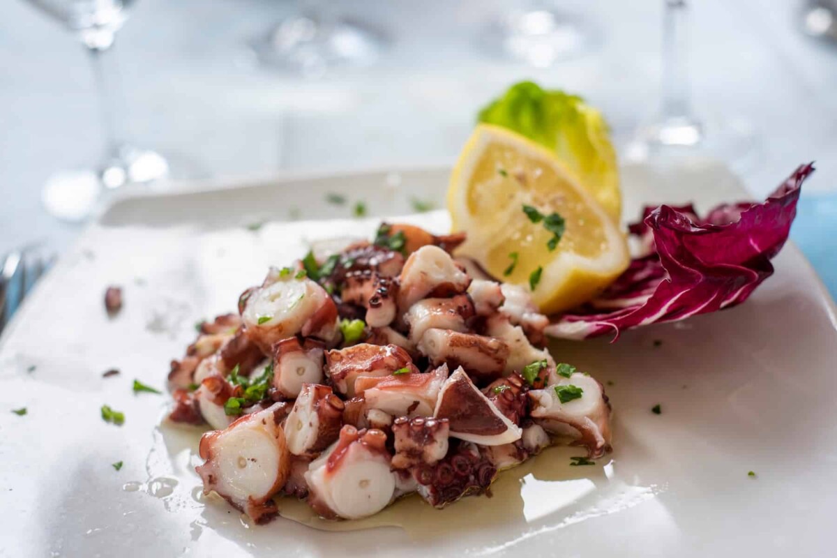 Salata od hobotnice poslužena na bijelom tanjuru, prelivena maslinovim uljem, ukrašena limunom, radičem i svježim peršinom.