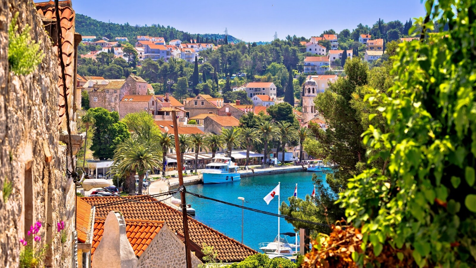 Cavtat, Konavle Pogled na Cavtat kroz uske kamene ulice, s crvenim krovovima, palmama uz rivu, brodicama u luci i zelenilom koje okružuje naselje, s crkvom i brdovitim krajolikom u pozadini.