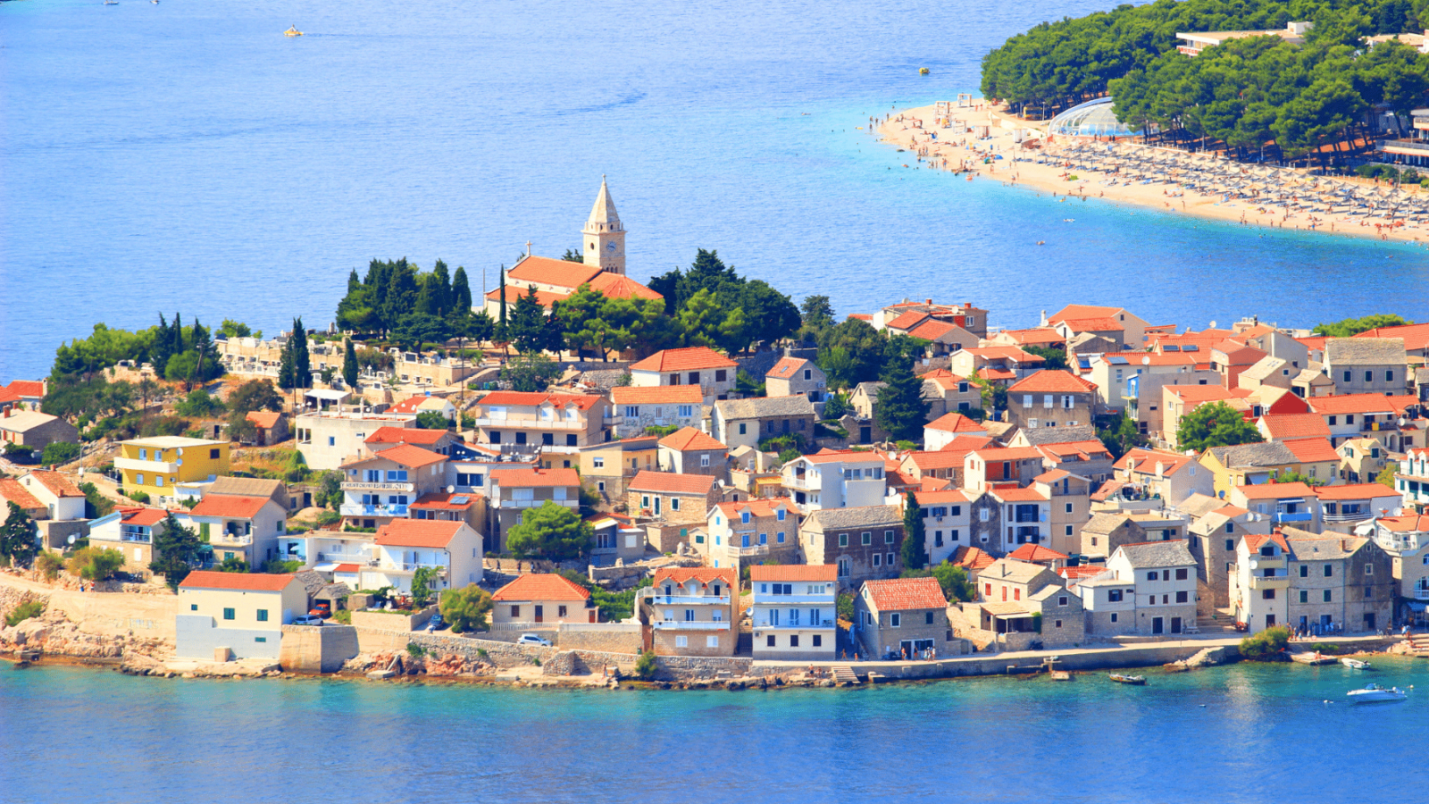 primosten-poluotok Primošten, slikovito dalmatinsko naselje uz more, s tradicionalnim kamenim kućama i crvenim krovovima, crkvom na uzvisini te popularnom plažom i borovom šumom u pozadini.