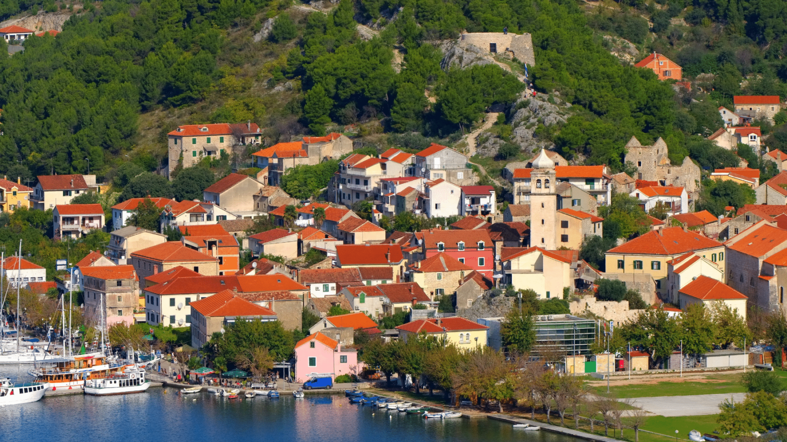 grad-skradin-dalmacija Pogled na Skradin – slikoviti dalmatinski gradić uz obalu, s crkvom, kamenim kućama s crvenim krovovima i zelenim brežuljcima u pozadini.