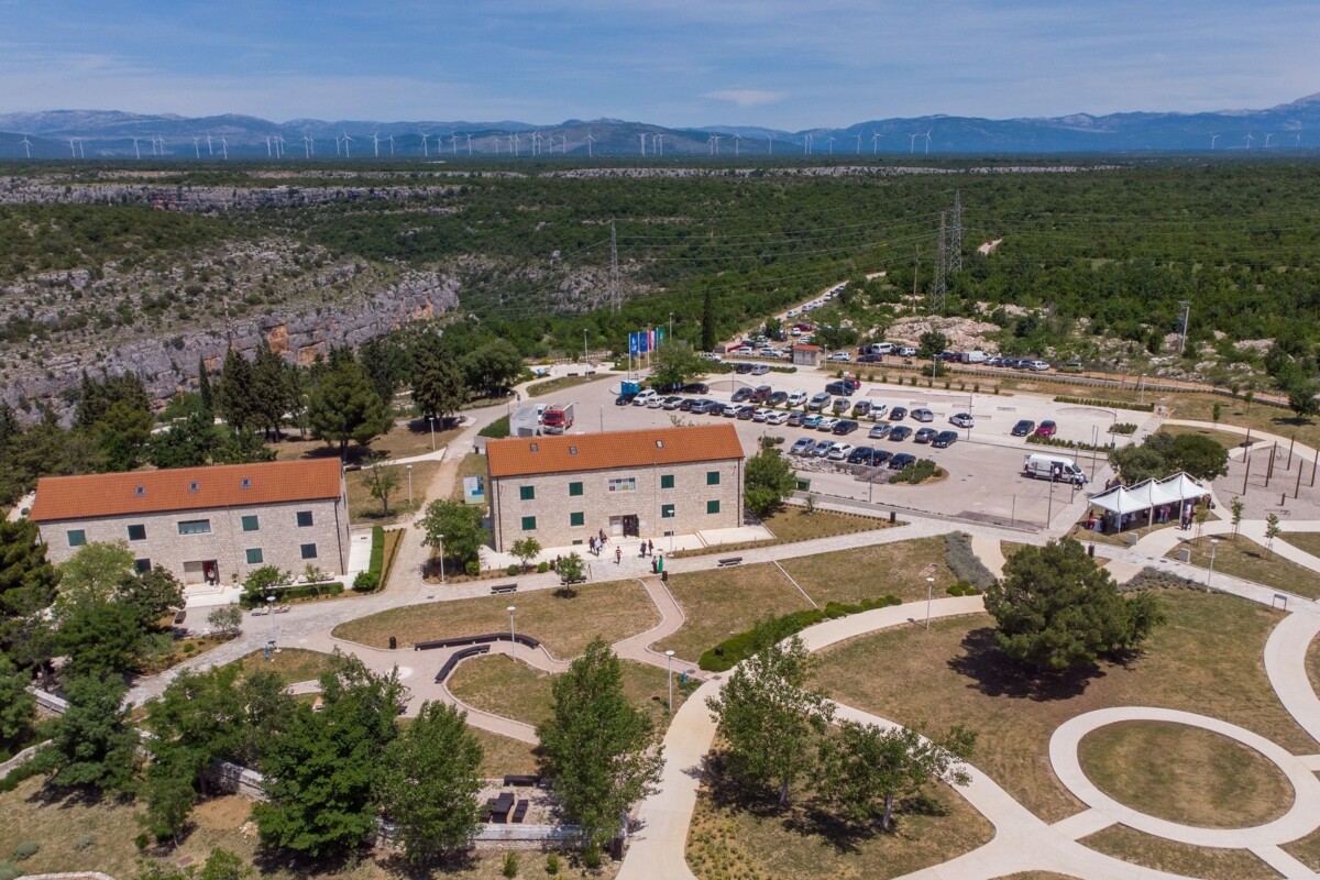 Eko-kampus-Krka Eko kampus Krka u Puljanima, moderno uređeni kompleks s kamenim zgradama, parkiralištem i uređenim šetnicama, smješten usred netaknute prirode Nacionalnog parka Krka, s pogledom na šumski krajolik i vjetroelektrane u daljini.