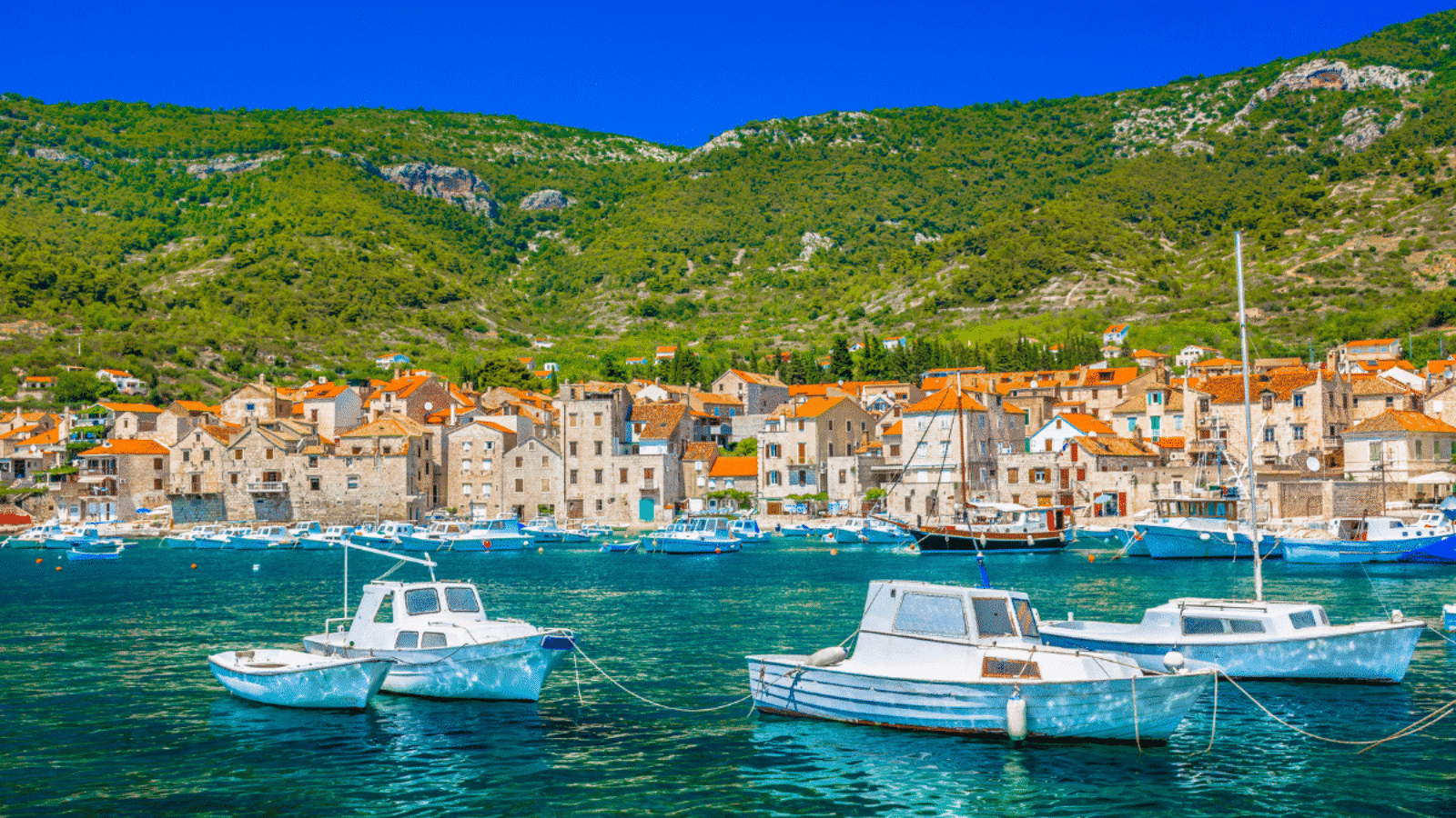 Pogled na Komišku luku na otoku Visu, s nizom bijelih brodica usidrenih u tirkiznom moru, starim kamenim kućama s crvenim krovovima uz obalu te zelenim brdima u pozadini.
