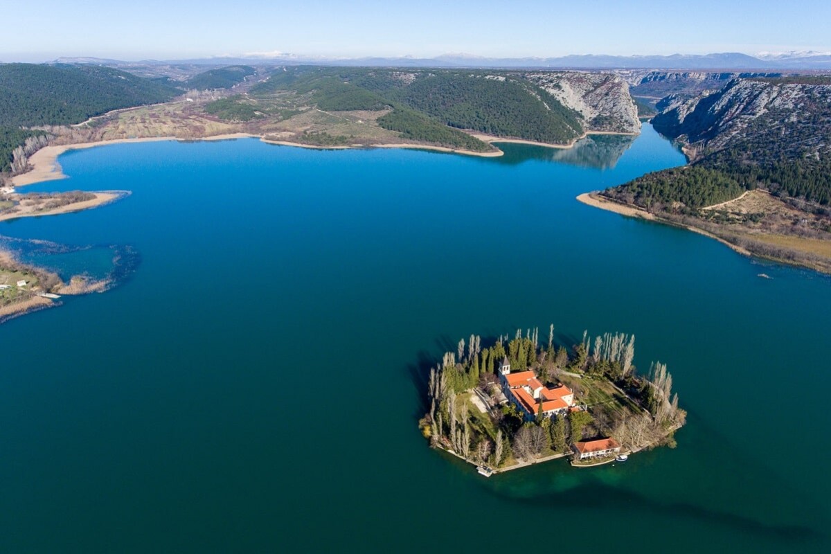 np-krka-otocic-visovac Otočić Visovac u Nacionalnom parku Krka, smješten usred modrozelenog jezera, s franjevačkim samostanom okruženim gustom vegetacijom, snimljen iz zraka uz pogled na okolne brežuljke i kanjon rijeke.