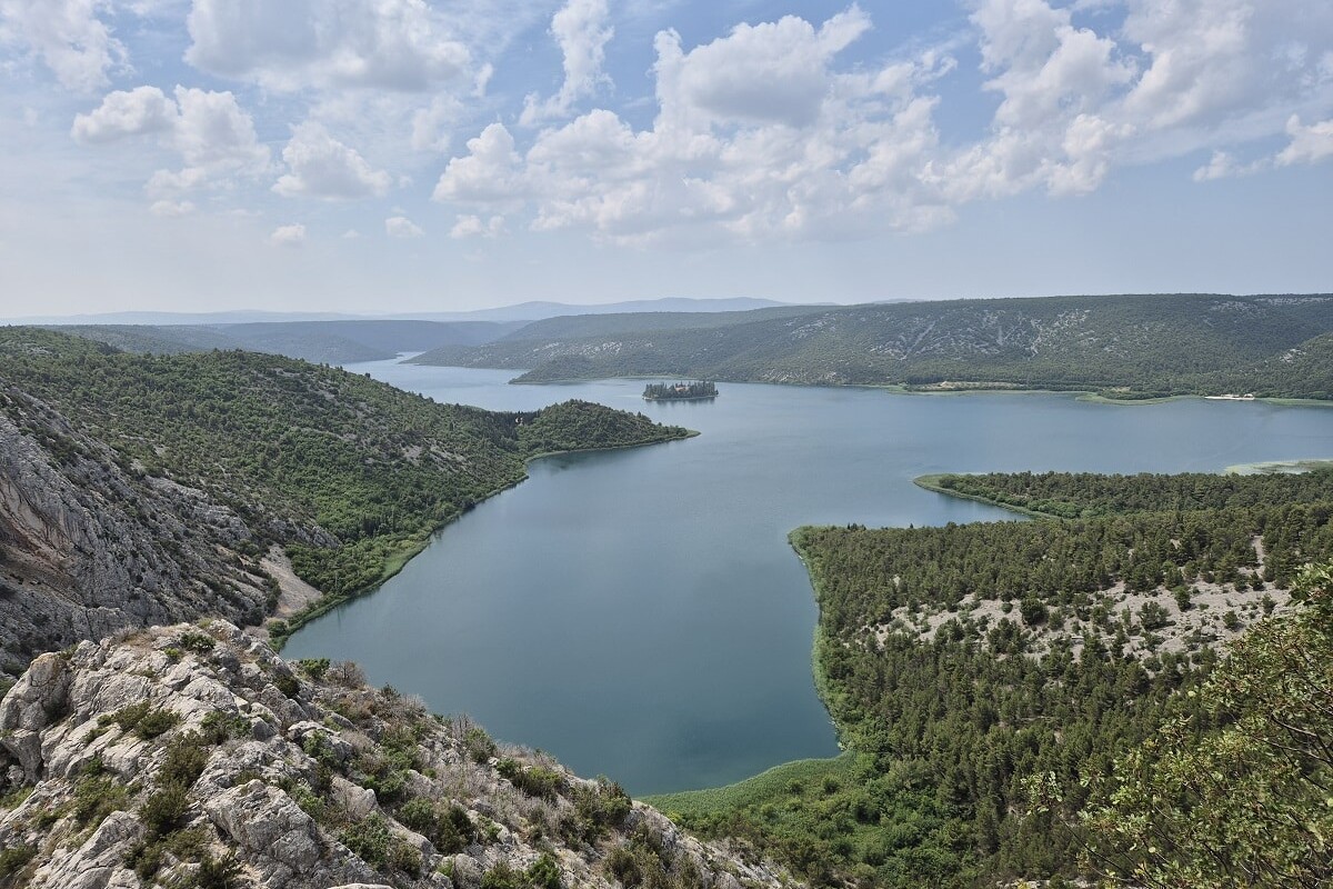 Pogled s vidikovca Rogovo u Nacionalnom parku Krka, koji pruža impresivan panoramski prizor na rijeku Krku okruženu zelenim brežuljcima, pod vedrim nebom s razbacanim oblacima.