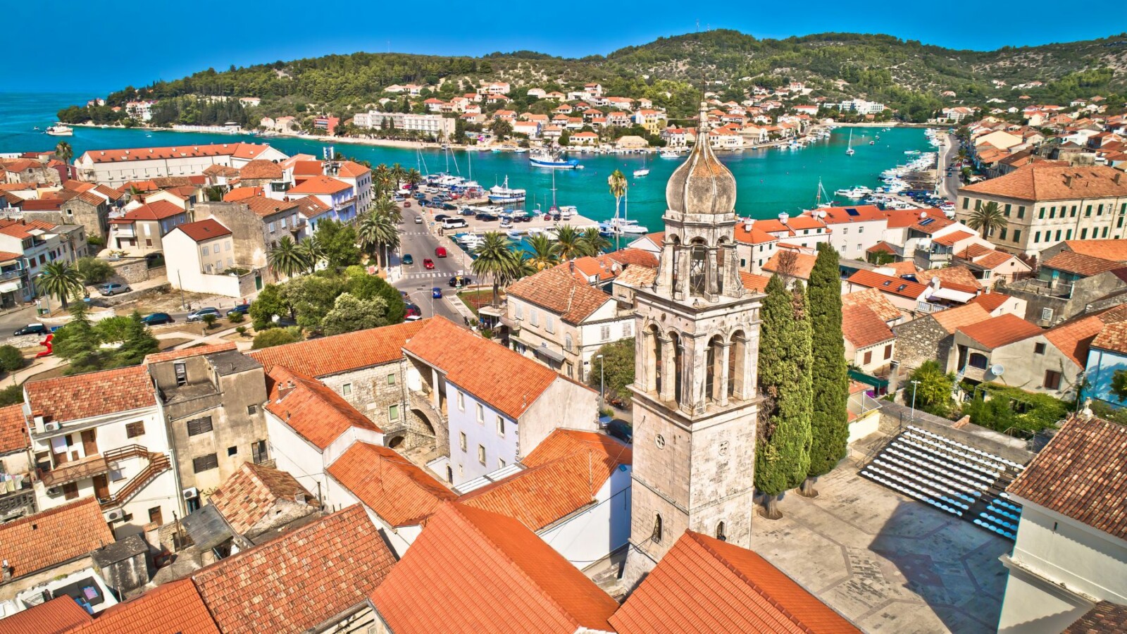 Pogled iz zraka na Vela Luku na zapadnom dijelu otoka Korčule, s istaknutom crkvom i zvonikom u središtu naselja, crvenim krovovima, uskim ulicama i prepoznatljivom uvalom s tirkiznim morem u pozadini.