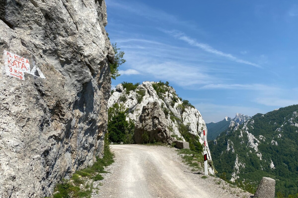Uska planinska cesta vijuga uz rub strmih stijena s pogledom na šumovite planine u daljini, a na stijeni uz cestu vidljiv je planinarski znak – Dabarska cesta i njezini impresivni krajolici.