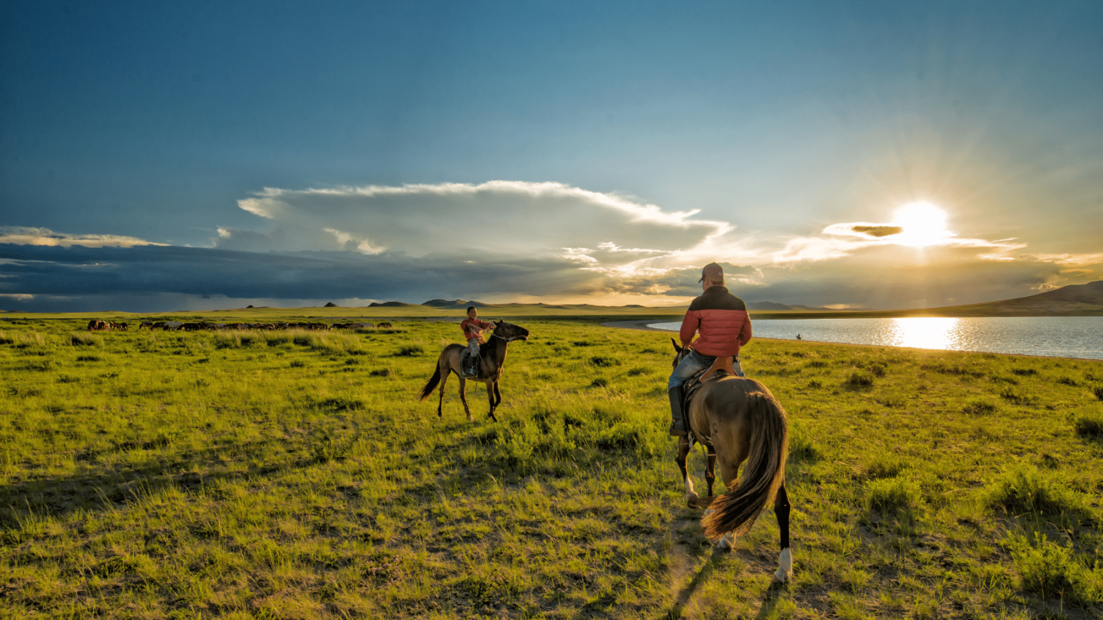 mongolija-priroda Mongolski jahači na konjima prelaze prostrane zelene stepe uz obalu jezera, dok sunce zalazi na horizontu, obasjavajući krajolik toplim zlatnim tonovima. Tradicionalni nomadski način života odražava se u ovom idiličnom prizoru mongolske prirode.
