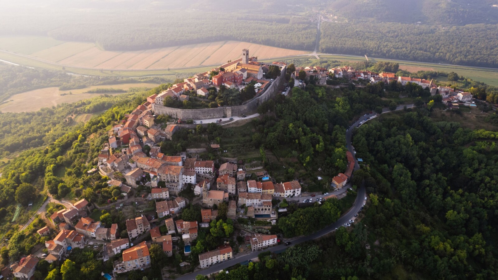 Pogled iz zraka na Motovun, srednjovjekovni gradić smješten na brežuljku, okružen gradskim zidinama i bujnim zelenilom, s poljima i šumama u daljini, koji čini jednu od najpoznatijih atrakcija u Istri.