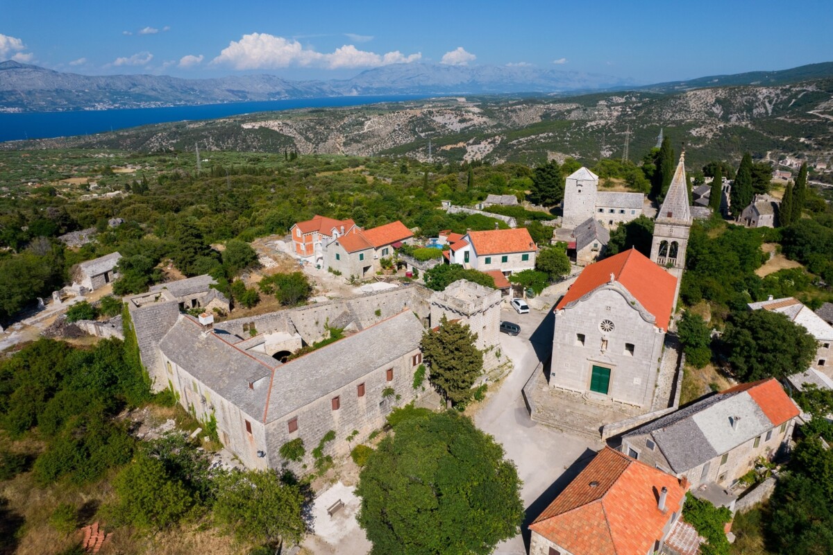 Pogled iz zraka na selo Škrip na otoku Braču s kamenim kućama, crkvom i crvenim krovovima, okruženo zelenilom i pogledom na more u daljini