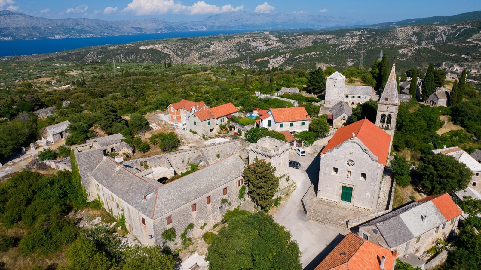 Pogled iz zraka na selo Škrip na otoku Braču s kamenim kućama, crkvom i crvenim krovovima, okruženo zelenilom i pogledom na more u daljini
