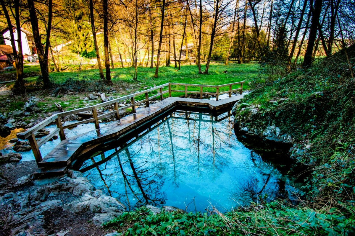 Termalni bazen Klevevž u Sloveniji – mali prirodni bazen s tirkiznom termalnom vodom, okružen drvenim mostićem i gustom šumom u pozadini.