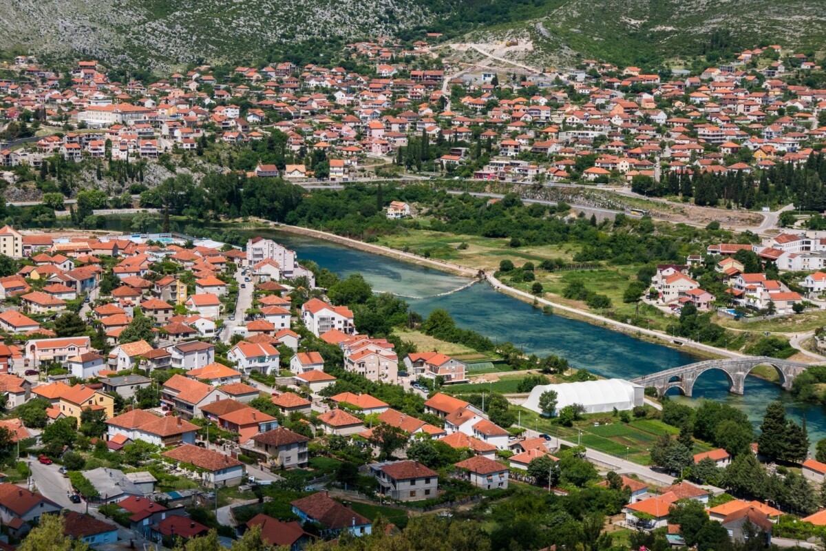 Pogled na Trebinje, grad u Republici Srpskoj, s rijekom Trebišnjicom i starim mostom, okružen kućama s crvenim krovovima.