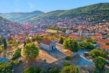 naselje-blato-otok-korcula Pogled iz zraka na naselje Blato na otoku Korčuli, s kamenitom crkvom smještenom na uzvisini usred zelenila, okruženom tradicionalnim dalmatinskim kućama i brdovitim krajolikom u pozadini.
