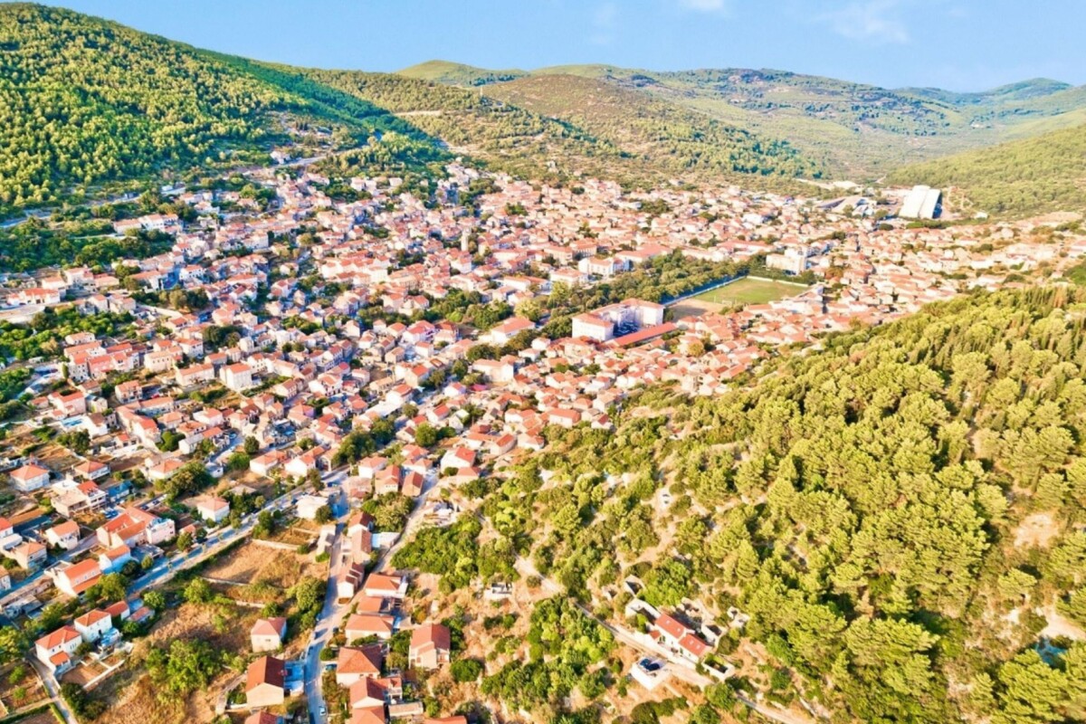 blato-korcula-panorama-iz-zraka Zračni prikaz naselja Blato na otoku Korčuli, smještenog između brda i prekrivenog gustom mrežom kuća s crvenim krovovima, uz prostrane šumske predjele i zelene padine koje ga okružuju.
