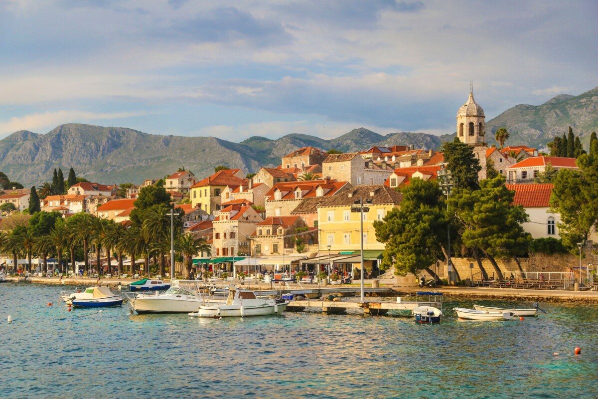 Cavtat, prekrasno priobalno naselje s kamenim kućama i crvenim krovovima, palmama uz rivu te brodicama u luci, s planinskim pejzažem u pozadini i zvonikom crkve koji dominira panoramom.
