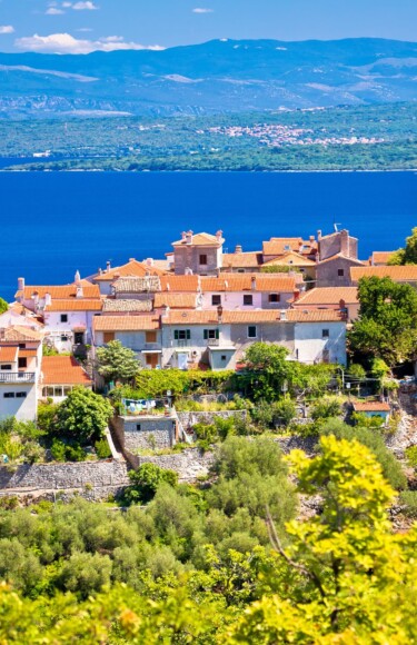 beli-otok-cres Naselje Beli na otoku Cresu, slikovito smješteno na uzvisini iznad Jadranskog mora, s kamenim kućama, crkvom i crvenim krovovima okruženima zelenilom, s pogledom na plavetnilo mora i kopno u daljini.