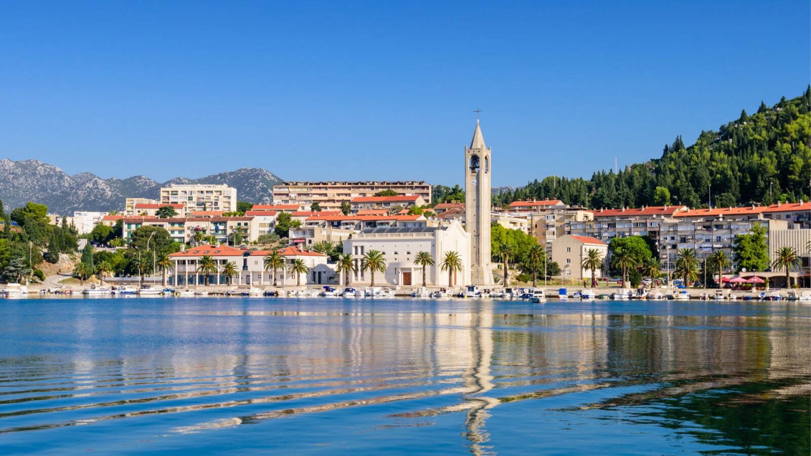 dolina-neretve-ploce Obalni dio grada Ploča u Dalmaciji s prepoznatljivom crkvom, refleksijom u mirnom moru i zelenim brdima u pozadini