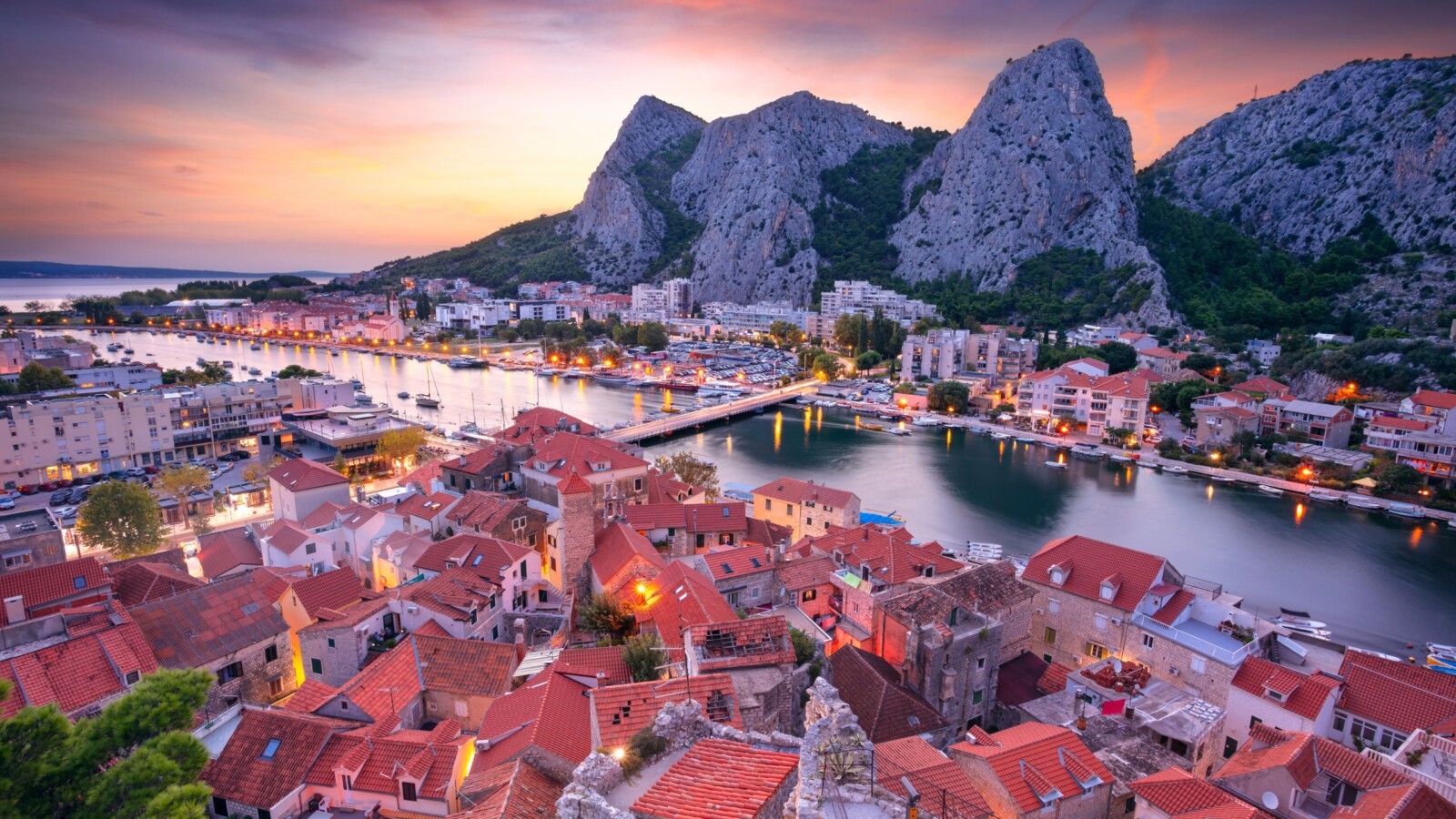 grad-omis-dalmacija Pogled na Omiš u suton – stari dio grada s crvenim krovovima uz rijeku Cetinu, okružen dramatičnim stijenama i osvijetljenim ulicama koje vode prema moru.