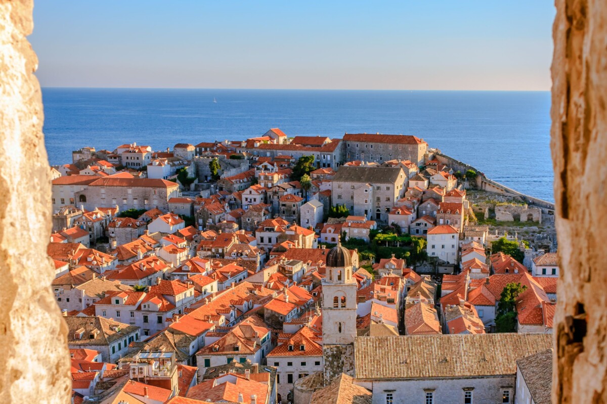 Pogled na staru gradsku jezgru Dubrovnika s kamenih zidina, s prepoznatljivim crvenim krovovima i plavetnilom mora u pozadini.