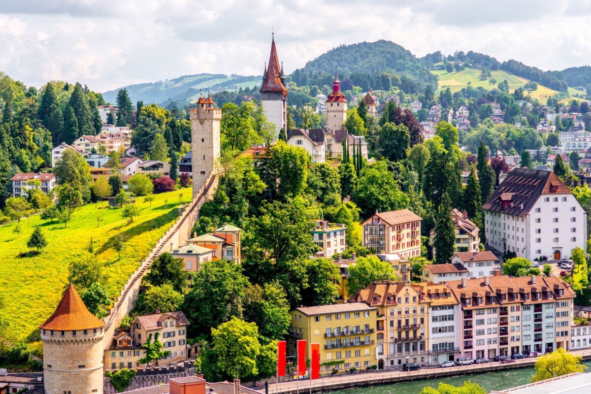 Pogled na slikoviti švicarski grad Luzern, s povijesnim zidinama, tornjevima i šarenim kućama uz jezero, okružen zelenim brežuljcima i planinama u pozadini.