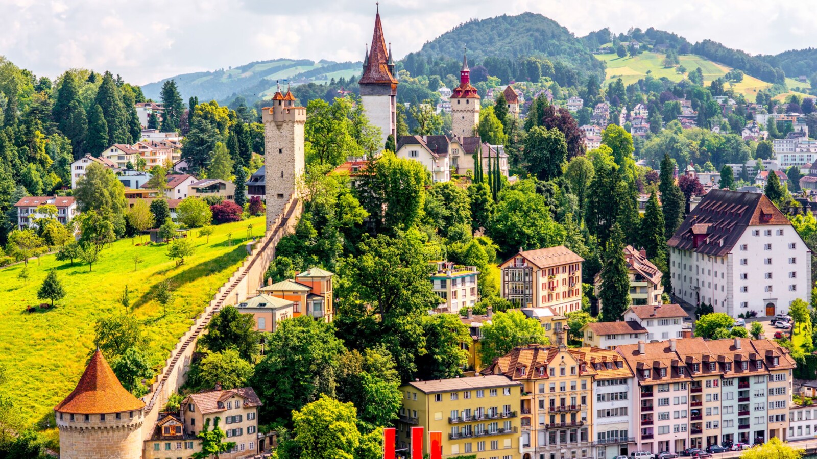 Pogled na slikoviti švicarski grad Luzern, s povijesnim zidinama, tornjevima i šarenim kućama uz jezero, okružen zelenim brežuljcima i planinama u pozadini.