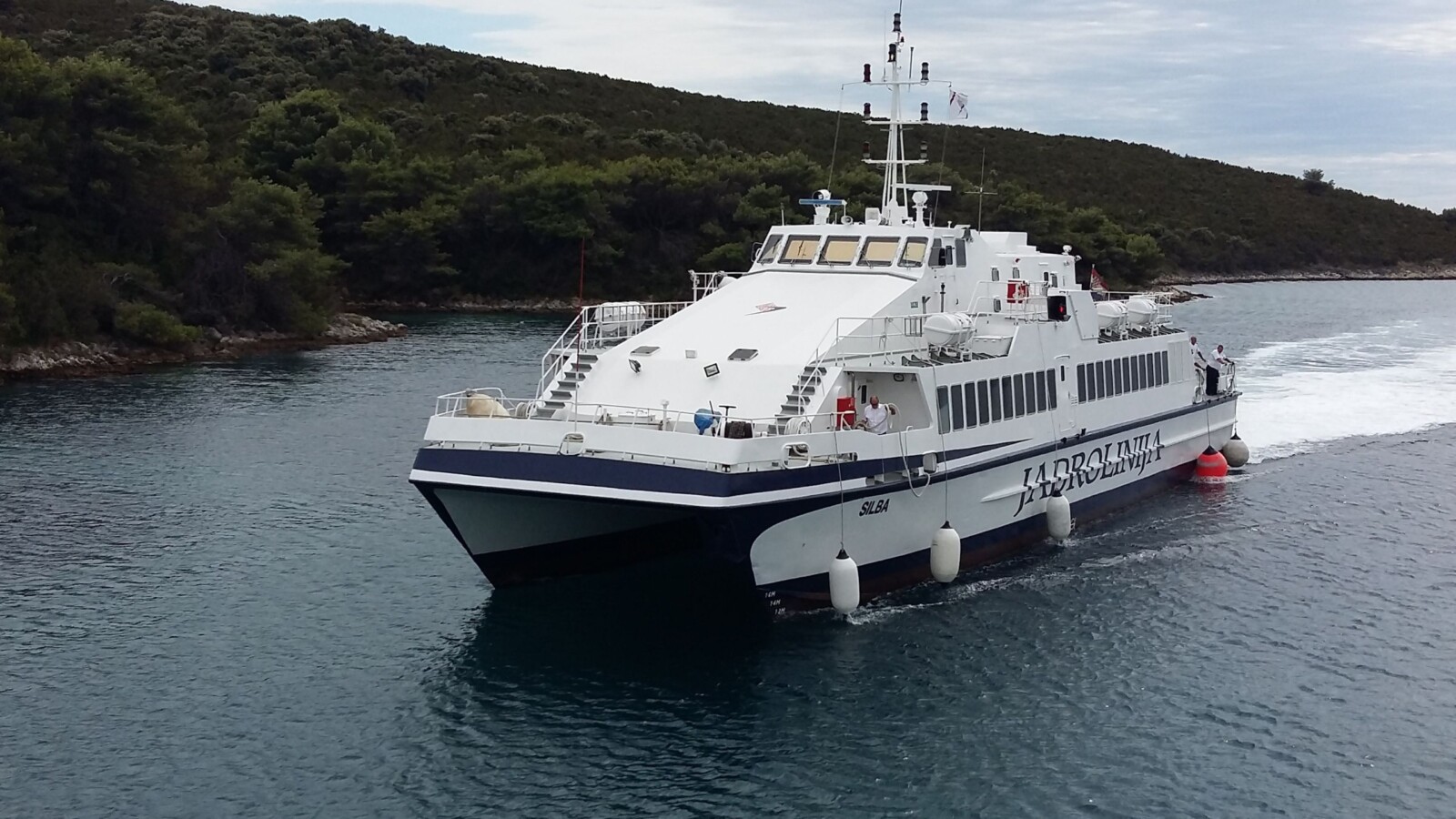 jadrolinija-trajekt Katamaran Jadrolinije na plovidbi kroz mirno more, okružen zelenilom obale.
