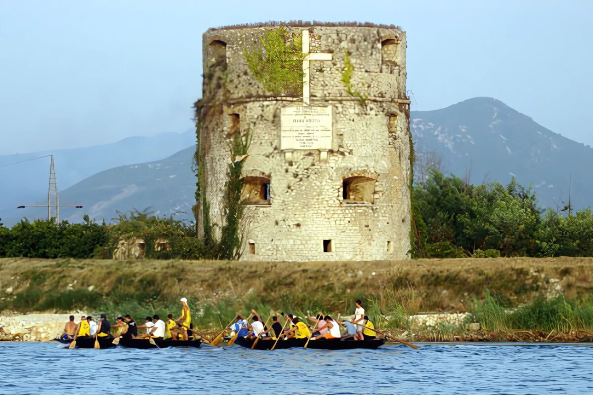 Kula Norinska u dolini Neretve, povijesna kamena utvrda s križem na fasadi, okružena prirodom i rijekom Neretvom, gdje veslači prolaze u tradicionalnim lađama tijekom maratona.