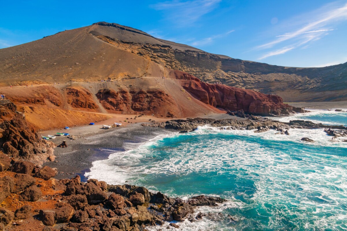 Pogled na vulkanski pejzaž Lanzarotea, s crvenkastim stijenama, crnom pješčanom plažom i valovima Atlantskog oceana koji udaraju o obalu.