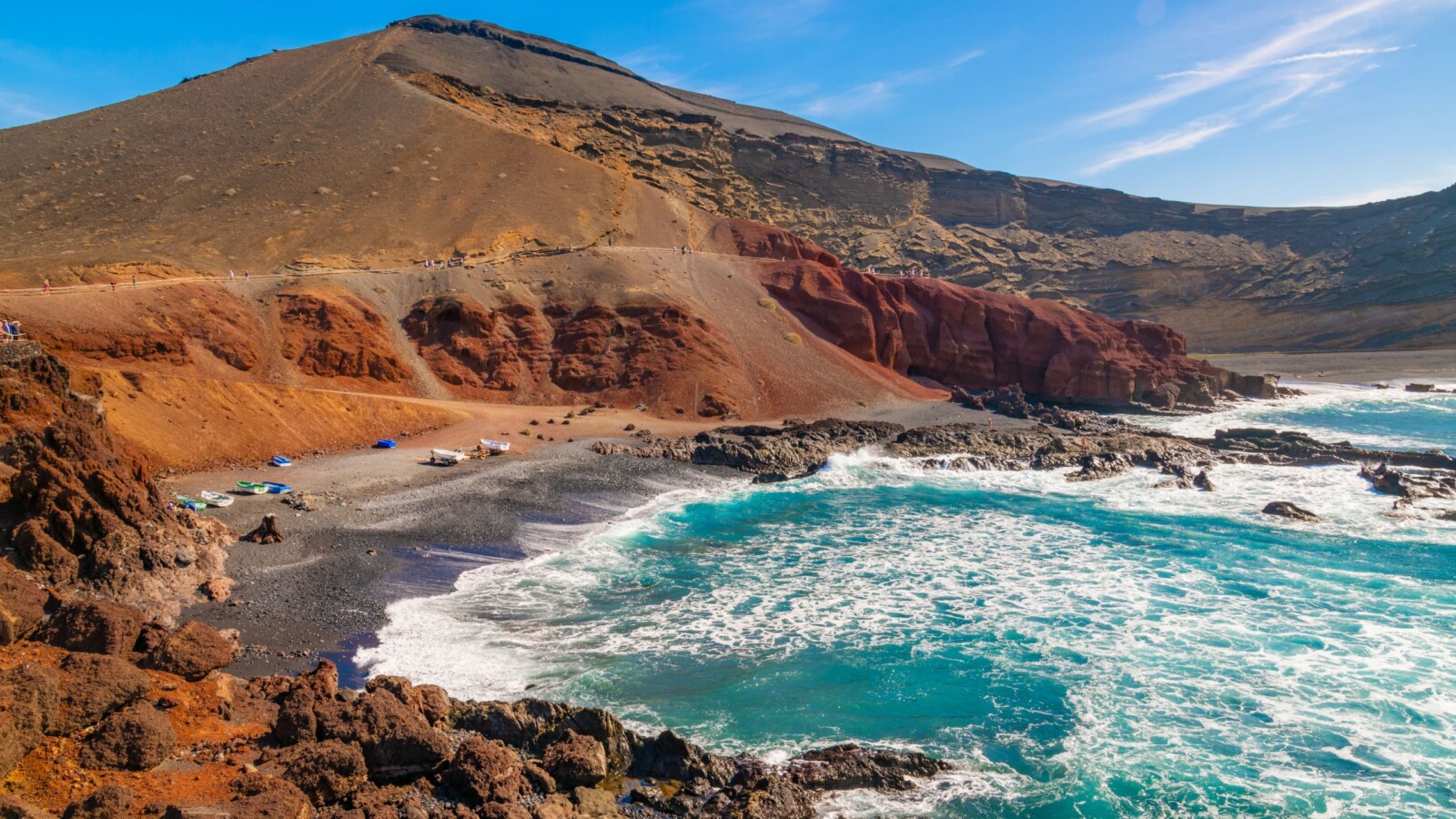 lanzarote-vulkanski-otok Pogled na vulkanski pejzaž Lanzarotea, s crvenkastim stijenama, crnom pješčanom plažom i valovima Atlantskog oceana koji udaraju o obalu.