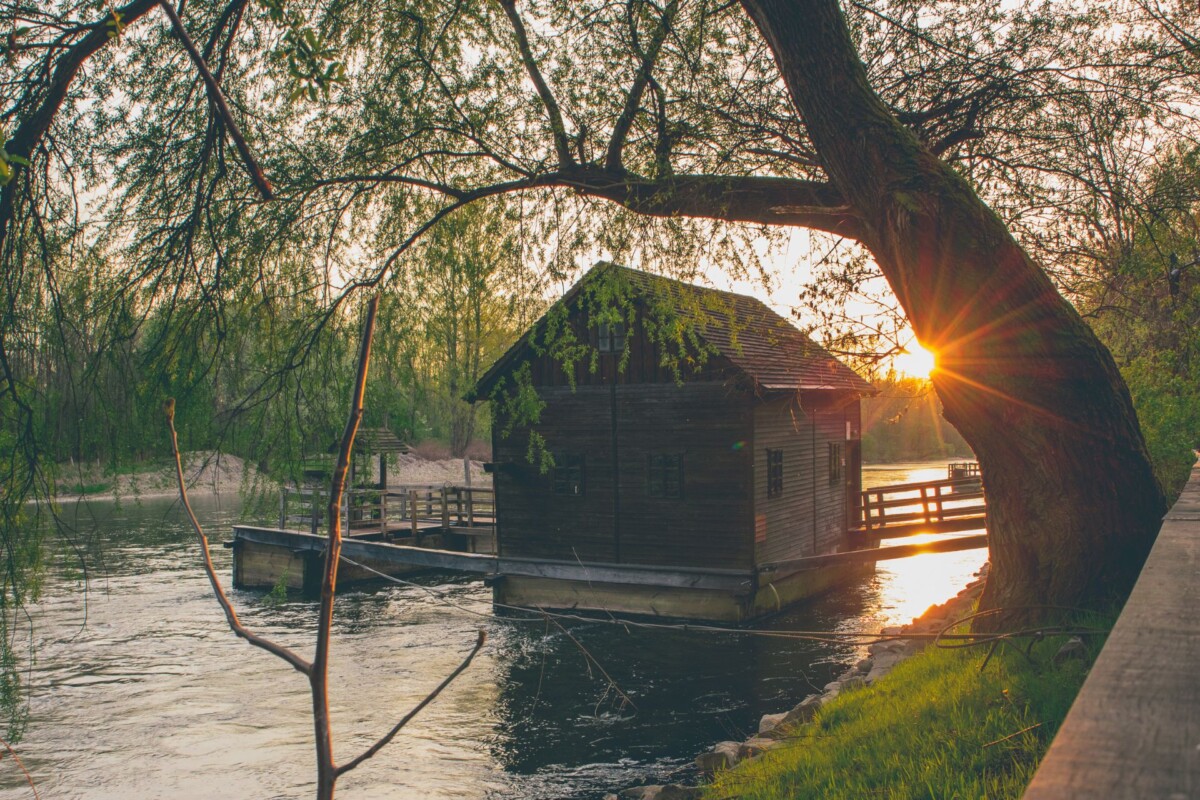 mlin-na-muri Drveni mlin na rijeci Muri u zalazak sunca, okružen zelenilom i refleksijom svjetla na vodi.