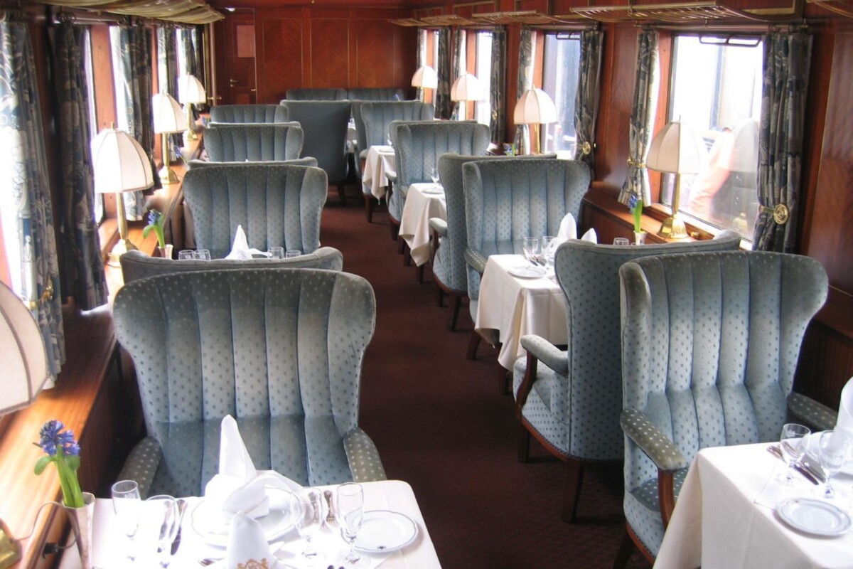 Unutrašnjost Orient Expressa s elegantnim plavim tapeciranim sjedalima i stolovima postavljenim za fine obroke, uz lampice na svakom stolu i drvene obloge na zidovima.