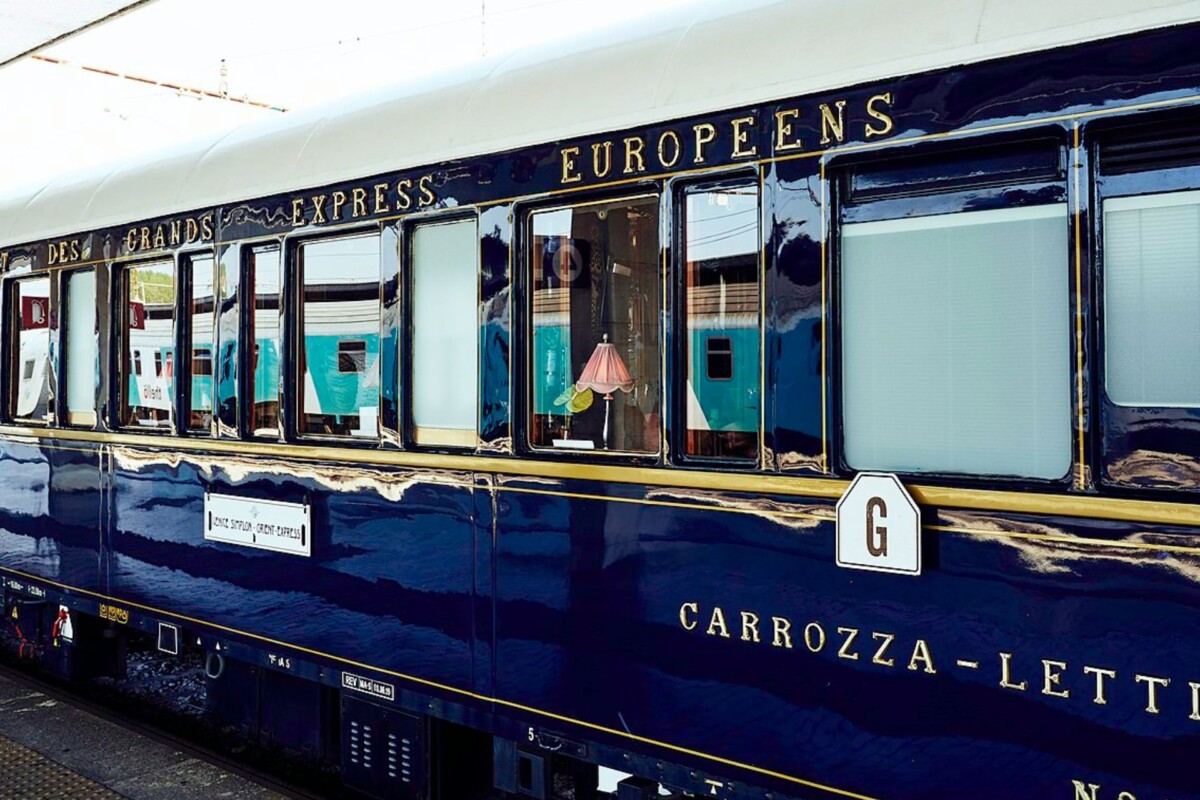 Detalj legendarnog Orient Expressa, luksuznog plavog vagona s natpisom 'Des Grands Express Européens' i prozorima kroz koje se vidi elegantna unutrašnjost s lampom.