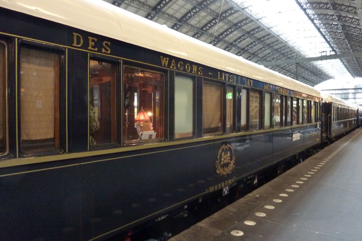 Vagon Orient Expressa na peronu, s tamnoplavo obojenom vanjštinom i zlatnim natpisima 'Wagons-Lits.' Kroz prozore se vidi luksuzno uređenje i diskretna svjetla unutrašnjosti.