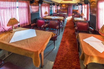 orient-express-vlak Unutrašnjost jednog od vagona Orient Expressa s vintage uređenjem, crvenim kožnim sjedalima, stolovima s bež stolnjacima i retro lampama, te tepihom u toplim tonovima.