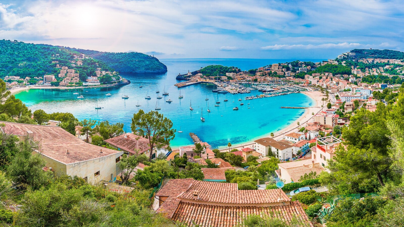 otok-palma-de-mallorca Pogled na slikovitu uvalu i grad Port de Sóller na Mallorci, s tirkiznim morem, brodicama u luci i zelenim brežuljcima okruženim mediteranskom arhitekturom.
