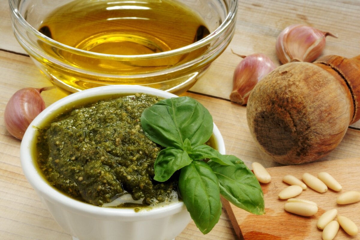 Svježe pripremljeni pesto alla Genovese u posudici s listom bosiljka, uz maslinovo ulje, češnjak, pinjole i drveni tučak na drvenoj podlozi
