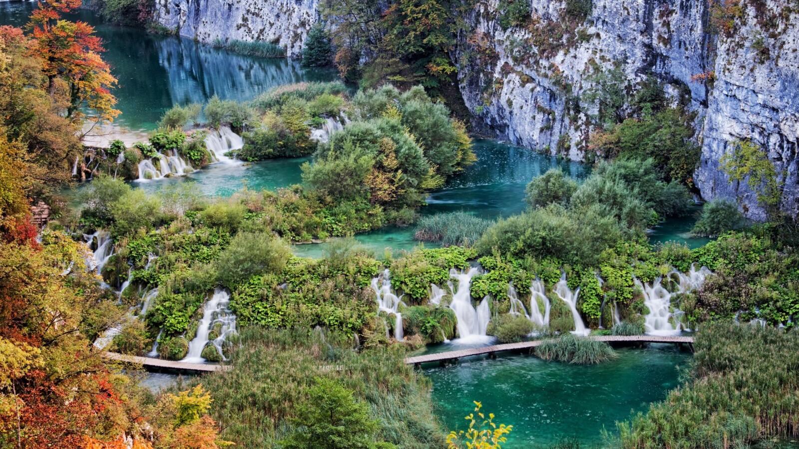 plitvicka-jezera-hrvatska Pogled na Plitvička jezera, slapovi i tirkizna voda okruženi zelenilom i stijenama, s drvenim stazama koje prolaze kroz bujnu vegetaciju u jesenskom ambijentu.