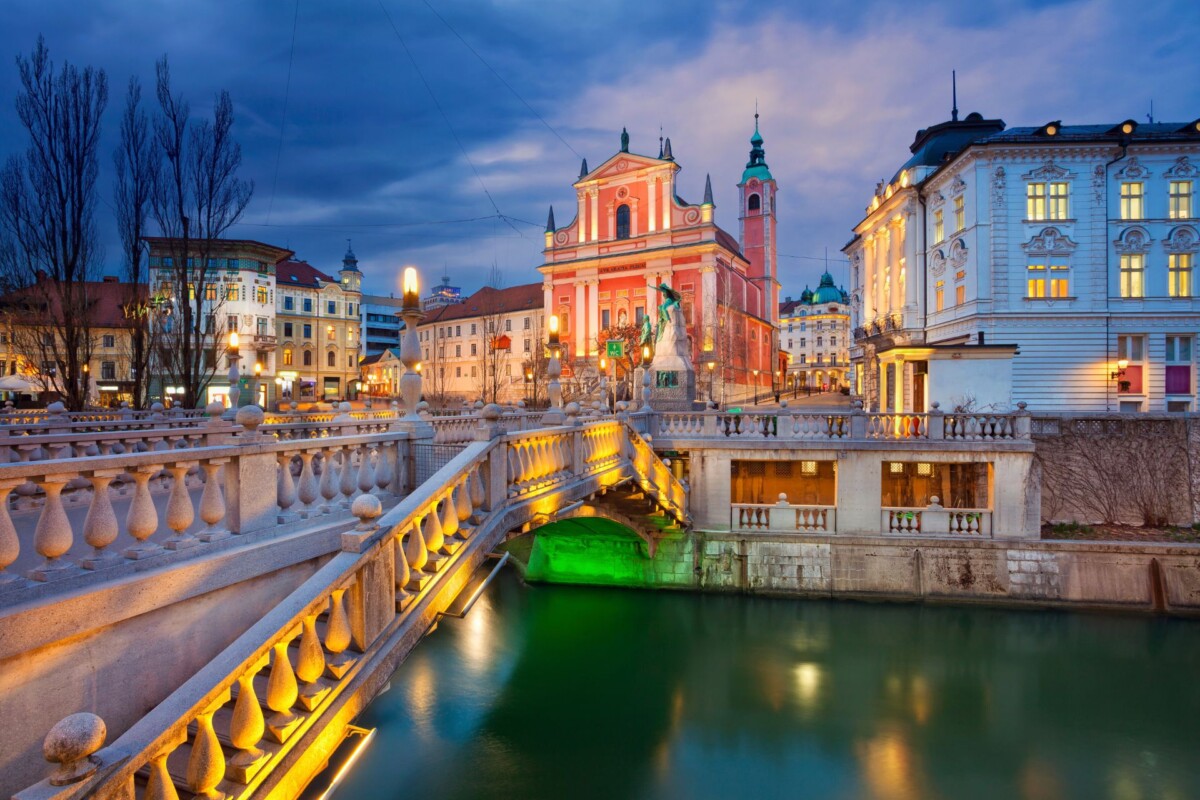 Tromostovje u Ljubljani u večernjim satima, s osvijetljenim mostovima i pogledom na Franjevačku crkvu u pozadini